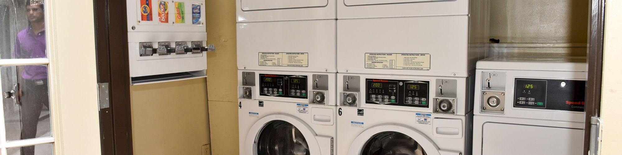 The image shows a small laundry room with two stacked washer-dryer units and a vending machine for soap, bleach, and softeners.