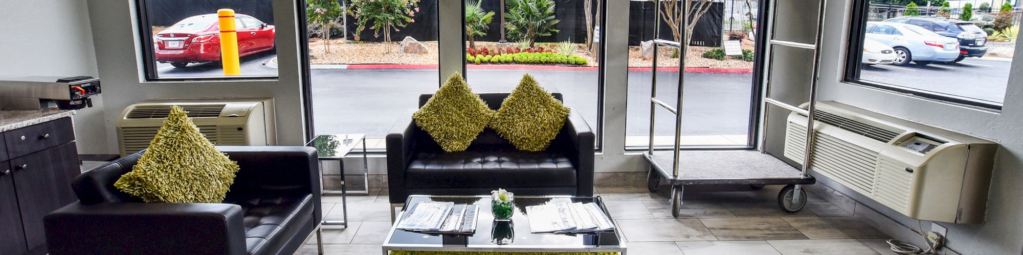 This image shows a small, modern lobby area with black couches, green pillows, a glass coffee table, and large windows overlooking a parking lot.