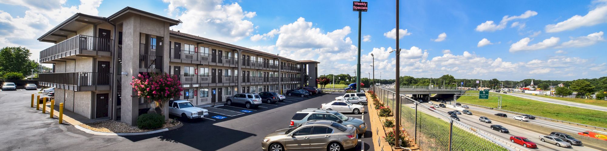 This image shows a hotel building, parking lot with cars, and a highway beside it under a clear blue sky with clouds, ending the sentence.