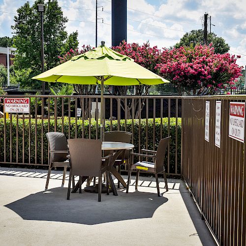 An outdoor seating area with a table, chairs, and a green umbrella, surrounded by a fence with pool rules and warning signs.