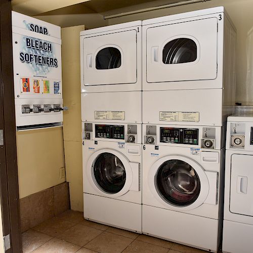 This image shows a small laundry room with stacked washers and dryers, a separate dryer, and a vending machine for soap, bleach, and softeners.