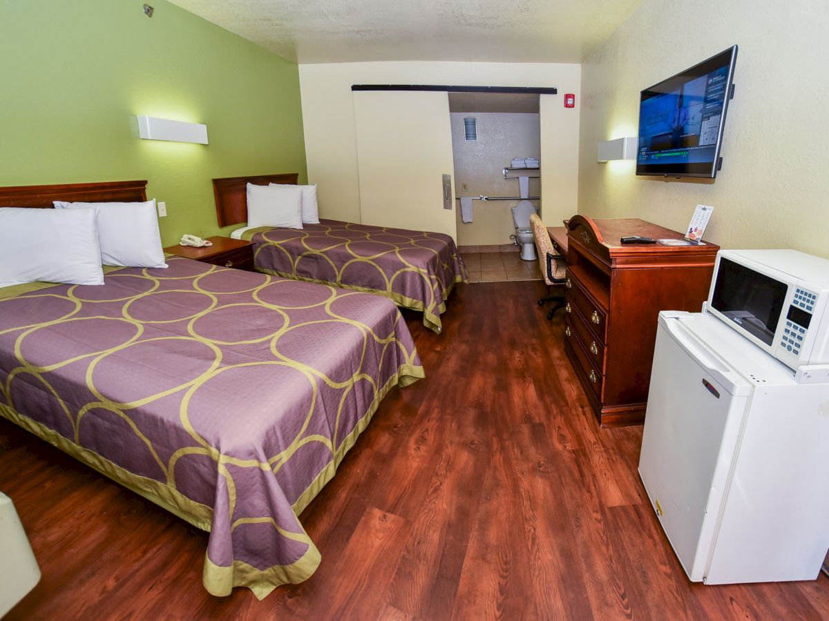 A hotel room features two beds, a TV, a white refrigerator with a microwave on top, wood flooring, and an ensuite bathroom in the background.