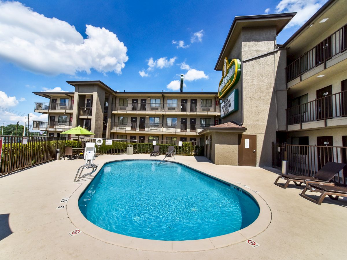 An outdoor pool in front of a multi-story hotel building, with lounge chairs and umbrellas under a clear blue sky.