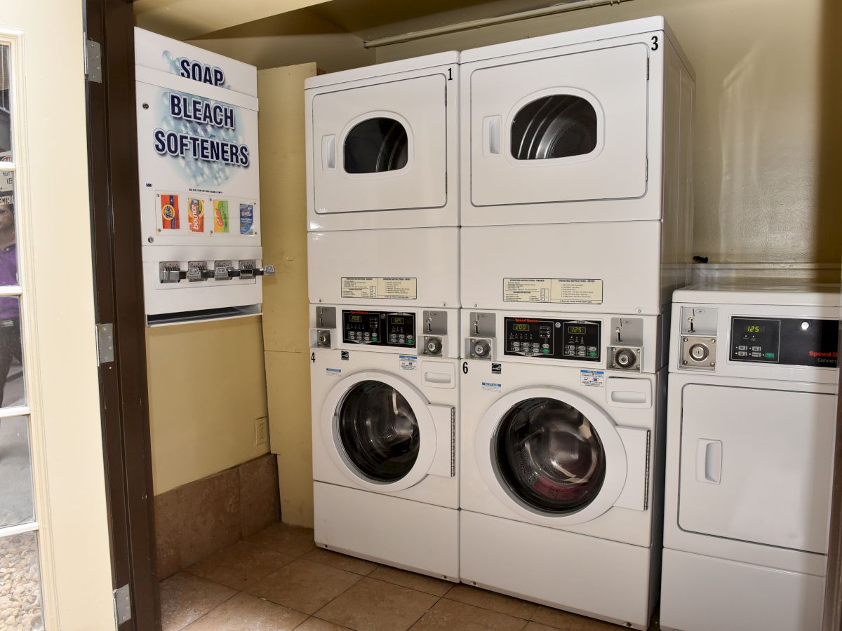 The image shows a small laundry room with two stacked washers and dryers, a small vending machine for laundry supplies, and a standalone dryer.