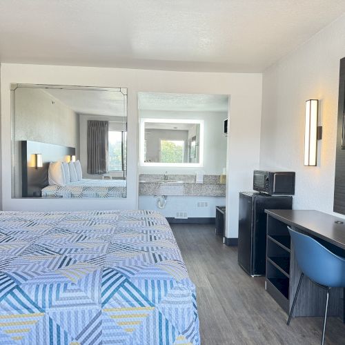 The image shows a modern hotel room with two beds, a desk with a chair, a TV, microwave, mini-fridge, and a large mirror reflecting a bathroom area.