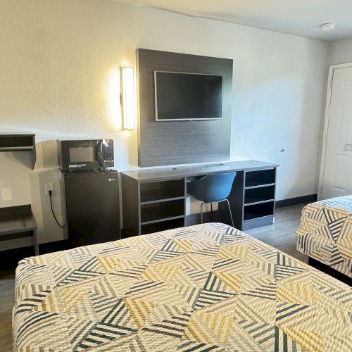 The image shows a hotel room with two beds, a desk and chair, a wall-mounted TV, a microwave, a mini-fridge, and a lamp on the wall.