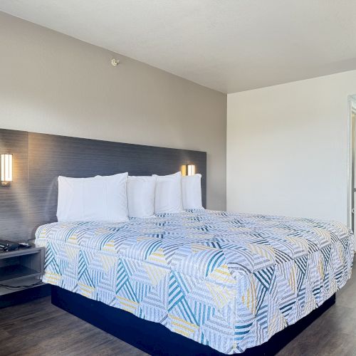 A tidy hotel room features a large bed with four pillows, modern wall lights, and an open bathroom doorway in the background.