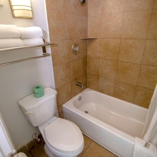 This image shows a small bathroom with a toilet, a stack of towels on a rack above it, and a bathtub with a shower curtain.