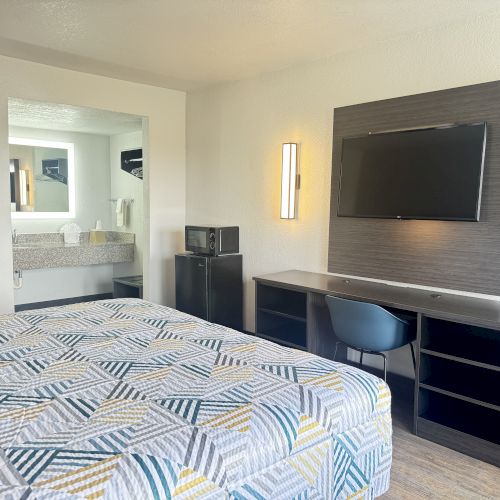 A modern hotel room with a bed, patterned bedspread, wall-mounted TV, desk, chair, microwave, and a visible bathroom sink and mirror area.