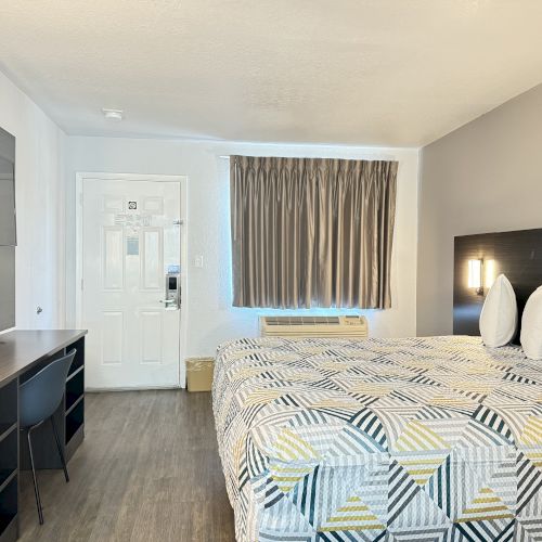 A hotel room with a neatly made bed, desk with chair, curtained window, wall-mounted TV, and a door. The decor is minimal and modern.