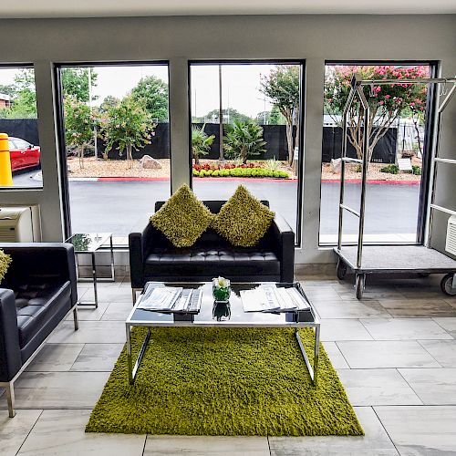 A cozy waiting area with black couches, green pillows, a coffee table, and a luggage cart, surrounded by large windows showcasing the outside view.