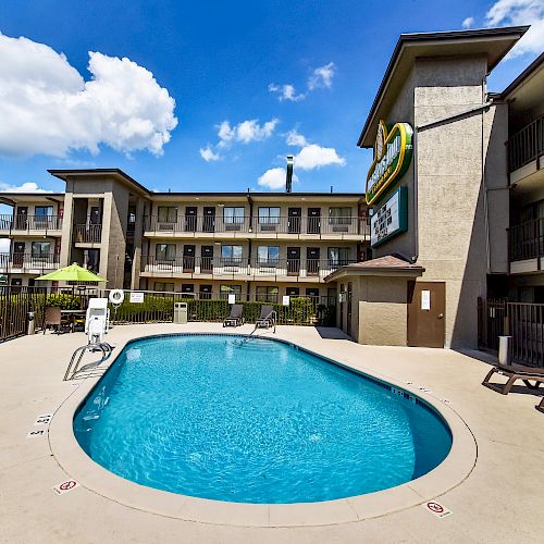 This image shows an outdoor swimming pool at a hotel with lounge chairs and a three-story building in the background under a clear blue sky.