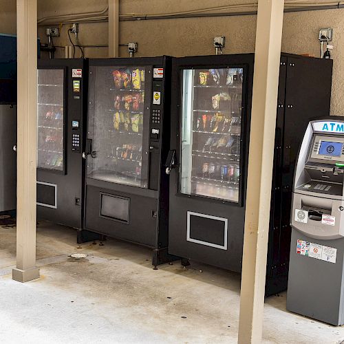 The image shows three vending machines and an ATM machine placed next to a washing machine and a trash bin in a sheltered area.