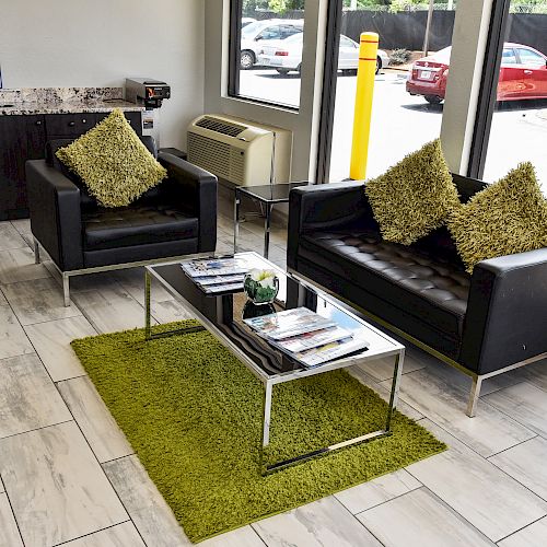 A waiting area with two black sofas, a glass coffee table on a green rug, green cushions, and magazines on the table by a window.