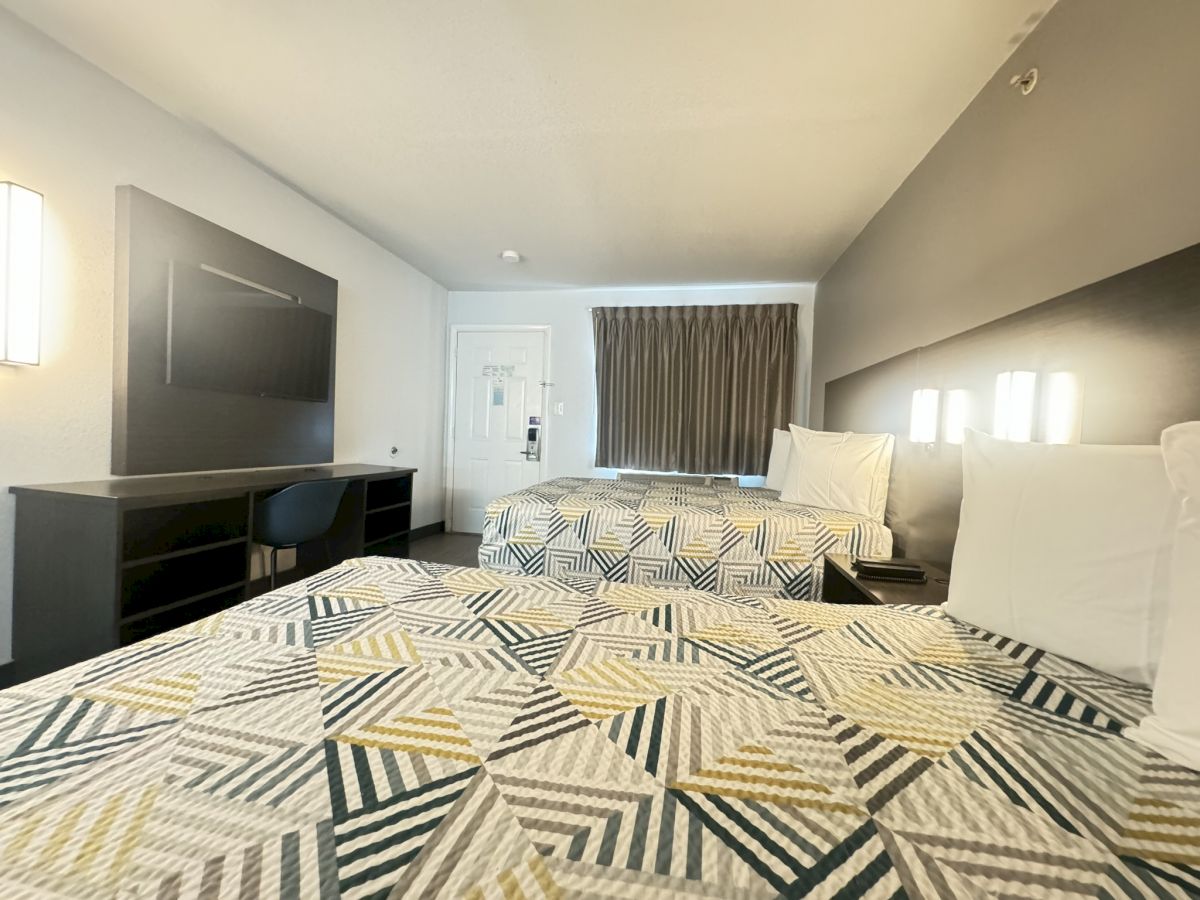 The image shows a hotel room with two beds, a desk, a TV stand, and a window with curtains. The decor features geometric patterned bedspreads.