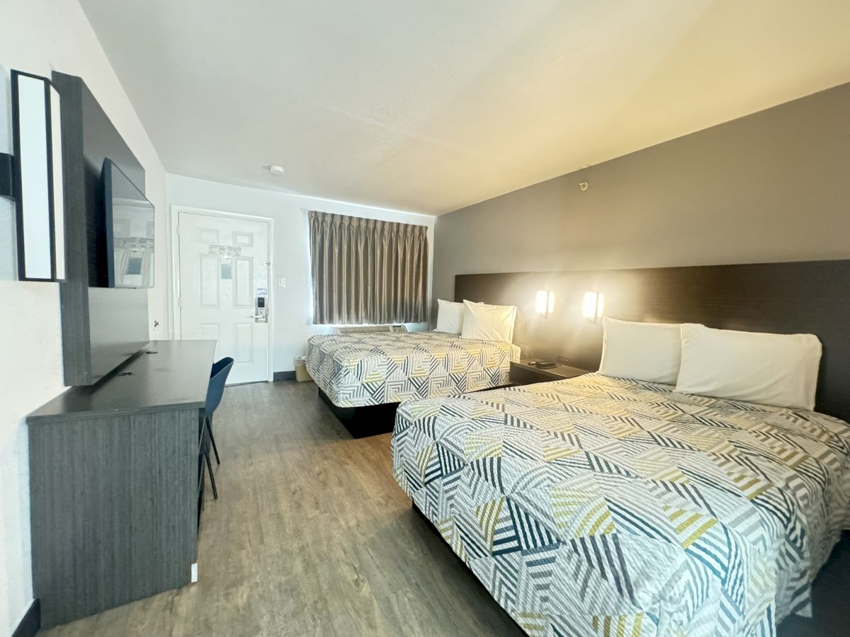 The image shows a clean, modern hotel room with two double beds, a desk and chair, a wall-mounted TV, wood-like flooring, and contemporary decor, ending the sentence.