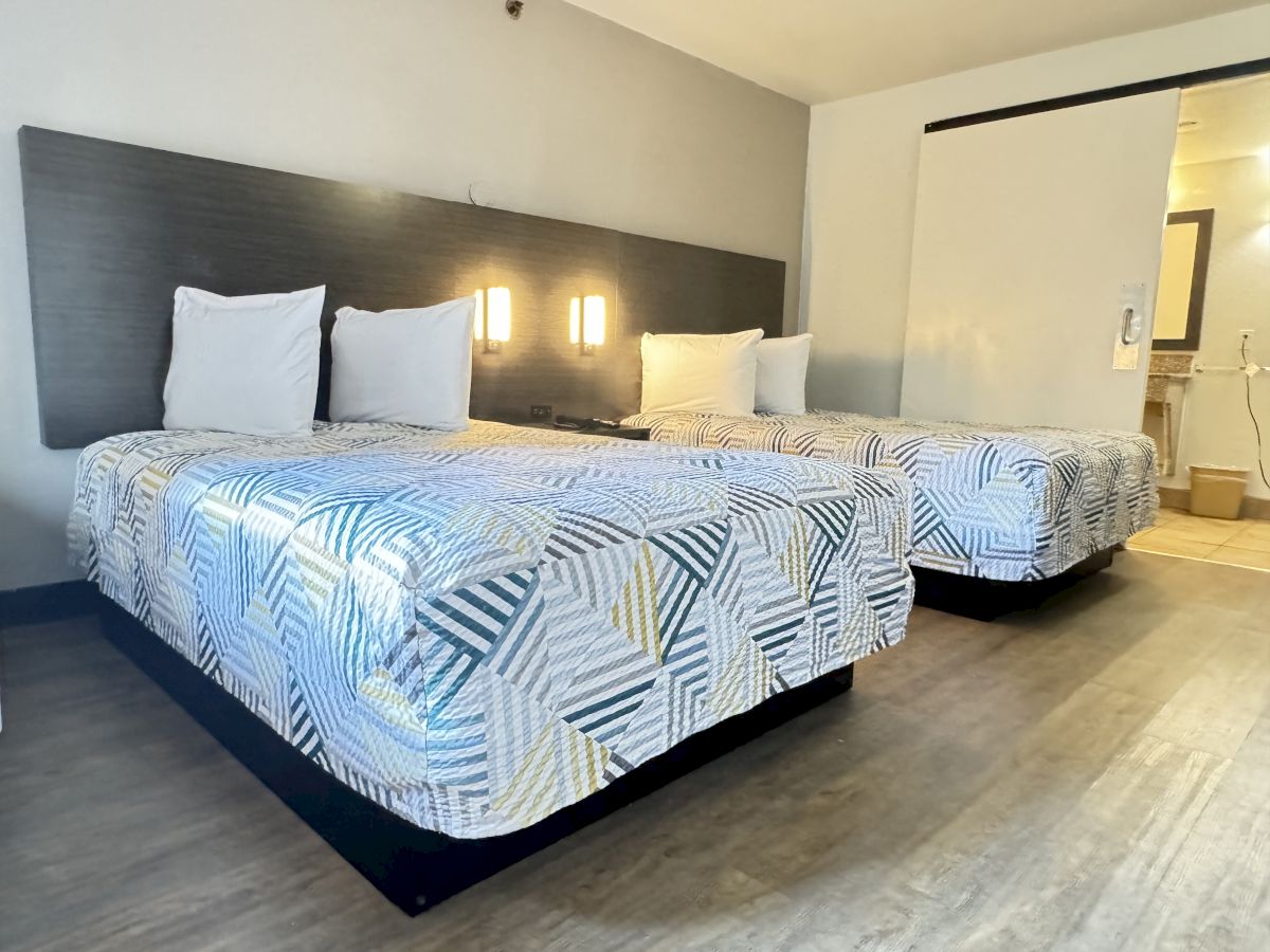 The image shows a modern hotel room with two double beds featuring patterned bedspreads, overhead lights, and a view of a bathroom in the background.