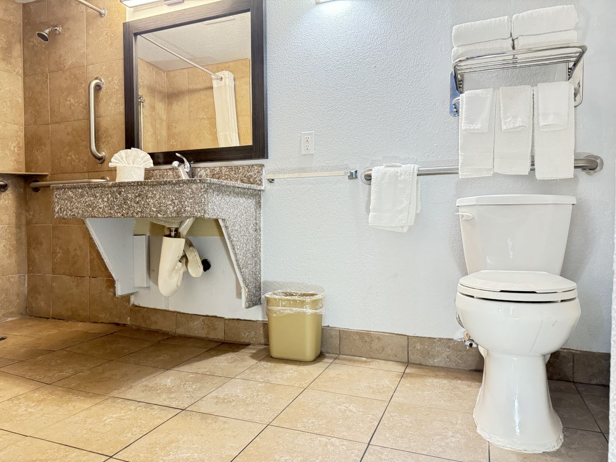 A bathroom with a toilet, sink, mirror, towel rack, trash bin, and neatly folded towels on a rail and shelf, all set in a tiled room.