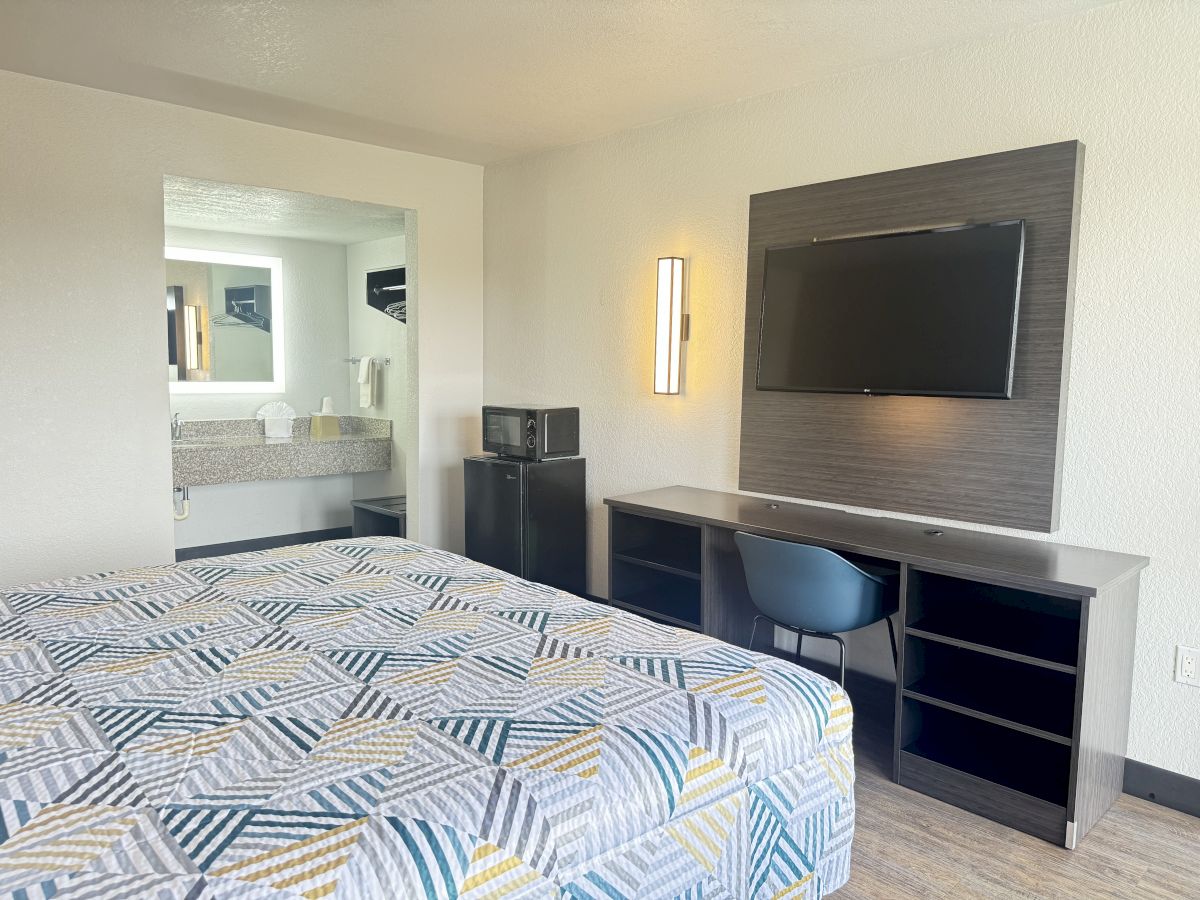 A neatly organized hotel room with a bed, wall-mounted TV, desk with chair, microwave, and a view into a bathroom area with a sink and mirror.