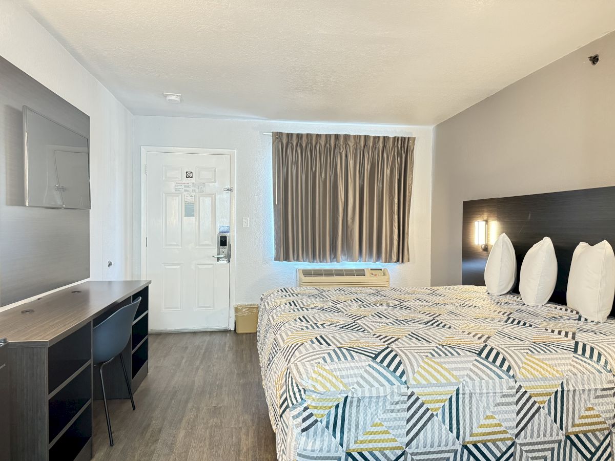 A neatly arranged hotel room with a large bed, desk, chair, TV, and curtains on the window. Decor is minimalist and modern.