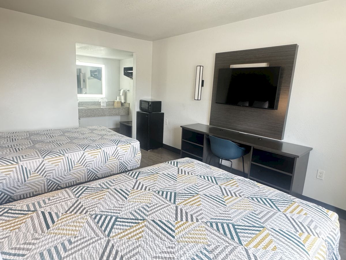 The image shows a hotel room with two beds, a TV mounted on the wall, a desk with a chair, and a small fridge next to a sink area in the background.