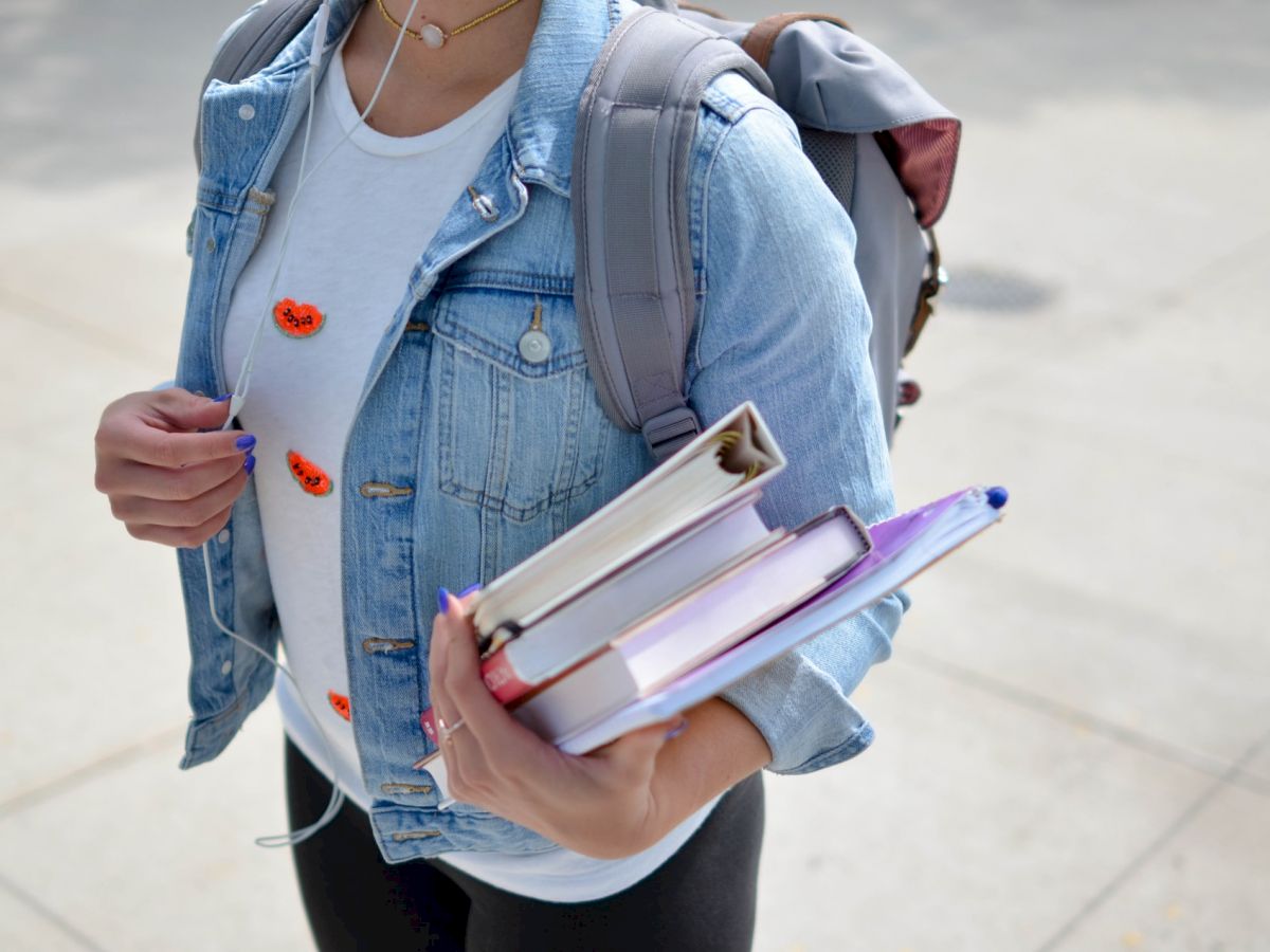 A person is carrying books and wearing a denim jacket with a backpack, standing on a paved surface.