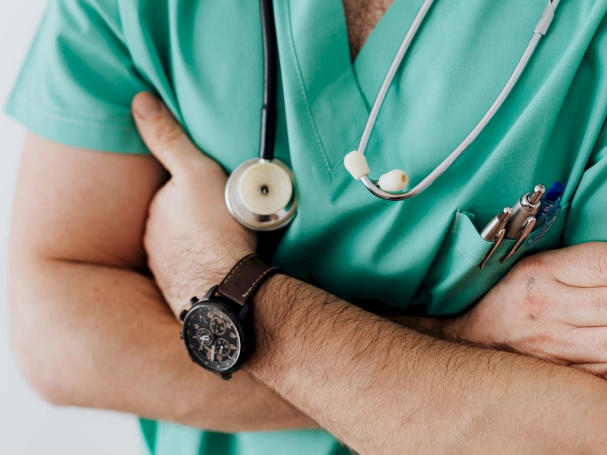 The image shows a person wearing green medical scrubs, a stethoscope, and a wristwatch, with arms crossed, and pens in the pocket.