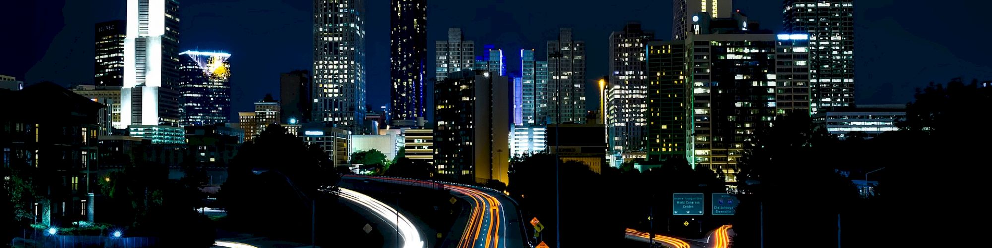 This image shows a bustling cityscape at night, with tall skyscrapers and glowing light trails from vehicles on the highways in the foreground.