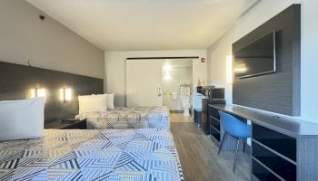 This image shows a hotel room with two double beds, a large flat-screen TV, a desk with a chair, and a bathroom visible in the back.