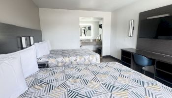 A modern hotel room features two beds with geometric-patterned bedspreads, a desk with a chair, a wall-mounted TV, and a view into the bathroom area.