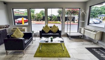 A modern waiting area with black sofas, green cushions and rug, a coffee table, and large windows. Luggage cart and air conditioner are visible.