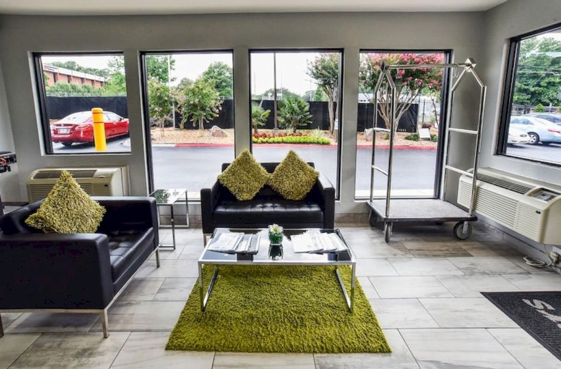 A modern waiting area with black sofas, green cushions and rug, a coffee table, and large windows. Luggage cart and air conditioner are visible.