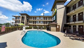 The image shows an outdoor swimming pool at a hotel or motel with a three-story building in the background and sunny weather.