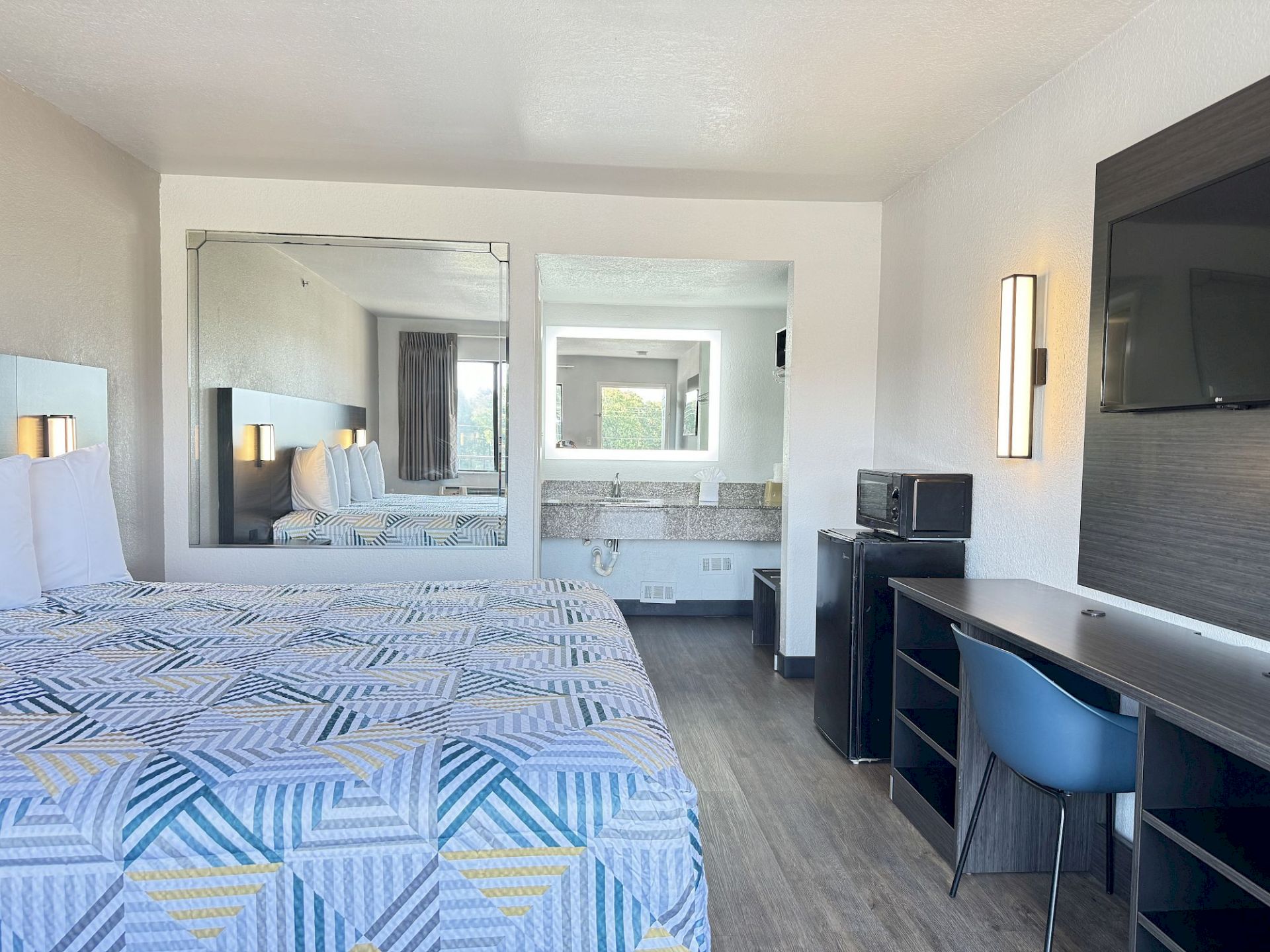 A hotel room with a bed, desk, TV, microwave, and a large mirror reflecting the room.