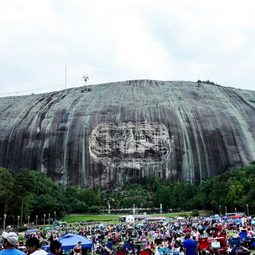 A large rock formation with a carved relief and a crowd of people gathered in a park area below, surrounded by trees under a cloudy sky.
