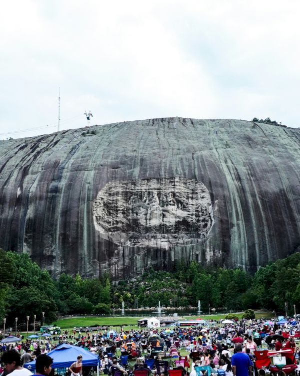 A large rock formation with a carved relief and a crowd of people gathered in a park area below, surrounded by trees under a cloudy sky.