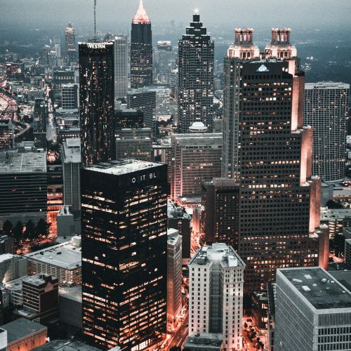 A cityscape with tall buildings during dusk, showcasing illuminated windows and streets, providing a modern urban atmosphere.