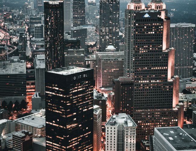 A cityscape with tall buildings during dusk, showcasing illuminated windows and streets, providing a modern urban atmosphere.