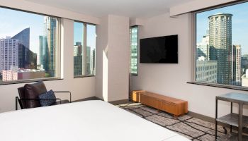 A modern room with a bed, TV, chair, windows showing a cityscape with tall buildings.