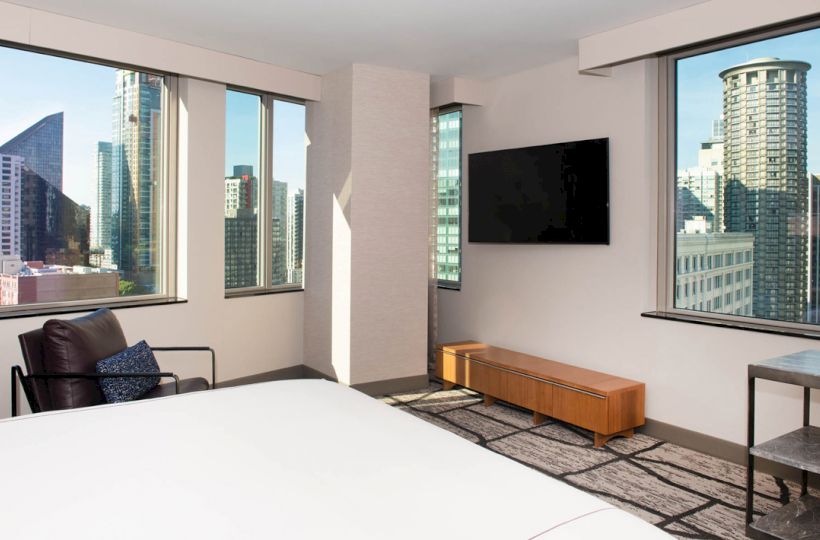 A modern room with a bed, TV, chair, windows showing a cityscape with tall buildings.