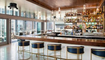 The image shows a modern bar with a wooden counter, high-top stools, and a wall of liquor bottles. Large windows offer a view of the cityscape.