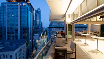 A modern rooftop terrace with seating areas and tables overlooking a cityscape with tall buildings, illuminated at dusk.