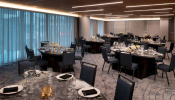 A banquet hall with round tables set for dining, each table has plates, glasses, and floral centerpieces. Blue curtains line the windows.