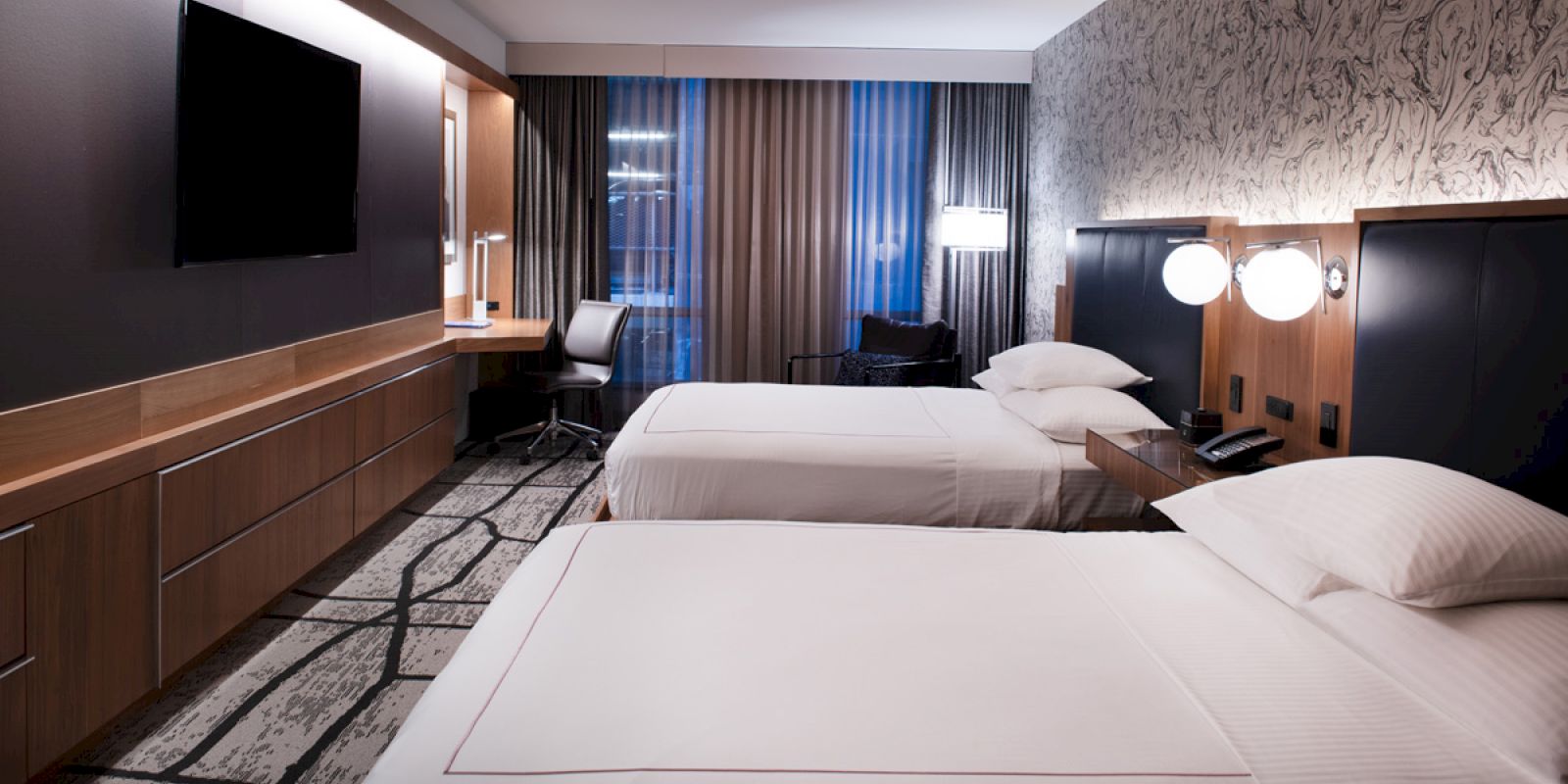 The image shows a hotel room with two beds, a large TV, a desk, and a chair in front of floor-to-ceiling windows with gray curtains.