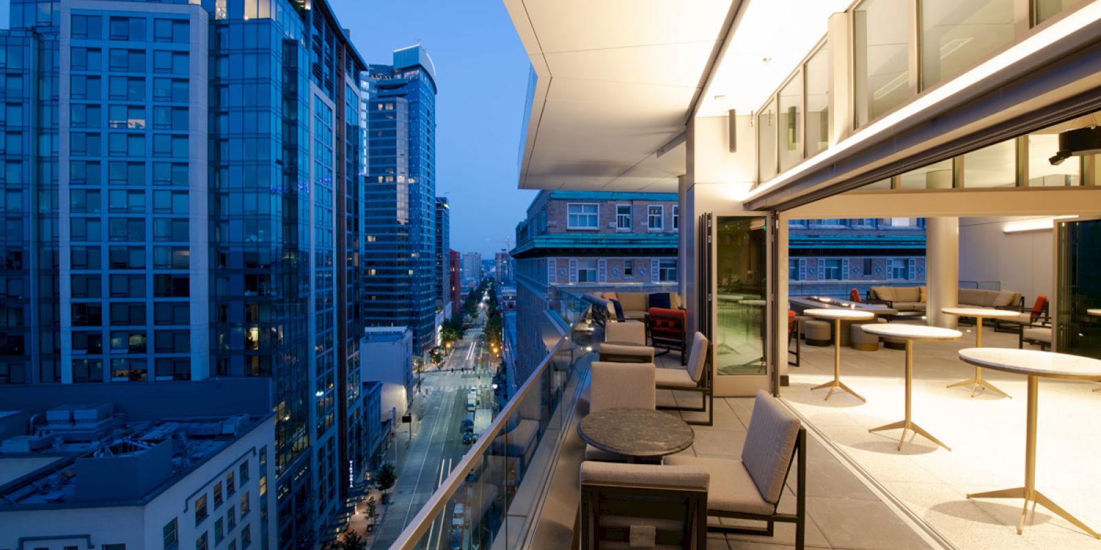 A modern cityscape view from a high-rise balcony, featuring outdoor seating and tables, with illuminated buildings lining the street below at dusk.