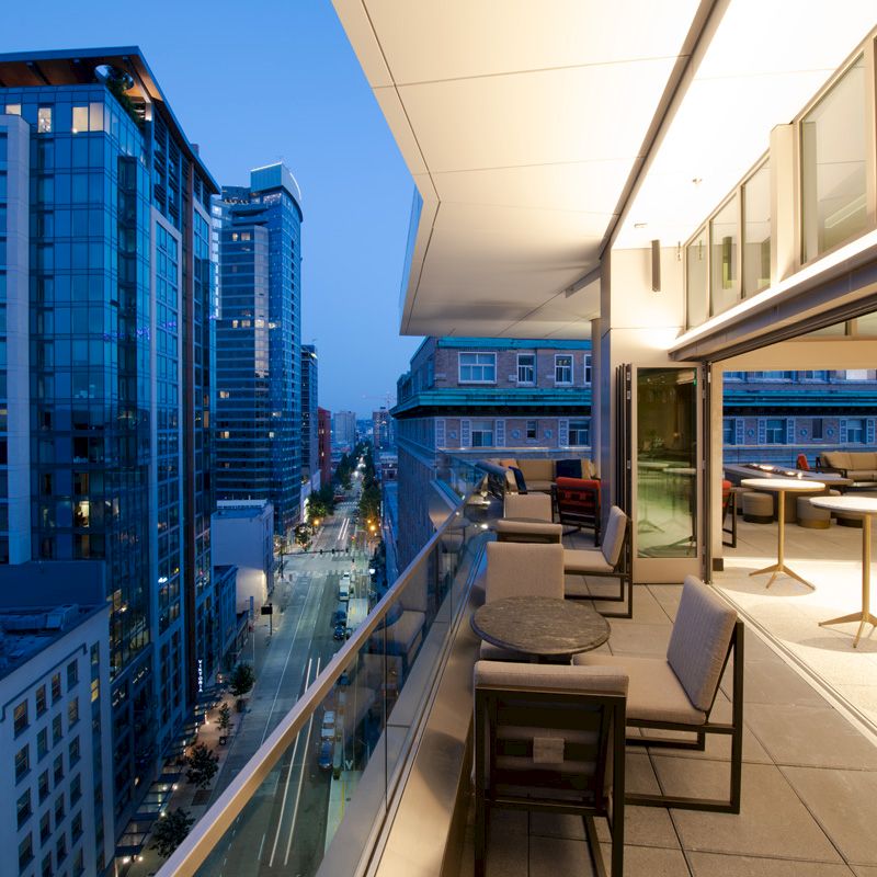 A modern urban balcony with seating overlooks a nighttime cityscape with tall buildings and a street below, featuring street lights and minimal traffic.