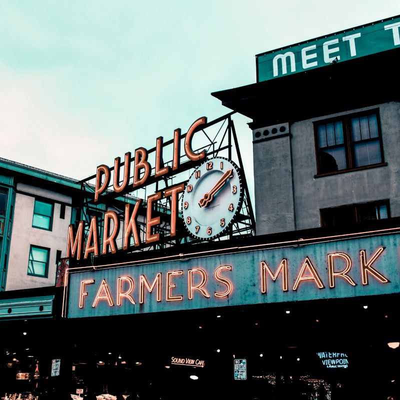 The image shows the signage of a public market with a neon sign reading "Farmers Market." A clock is displayed above the "Market" sign.