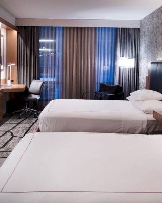 A modern hotel room with two double beds, a TV, a desk, and large windows with curtains, featuring a monochrome color scheme.