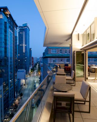 The image shows a modern cityscape in the evening, with tall buildings and a stylish balcony featuring outdoor seating and tables.