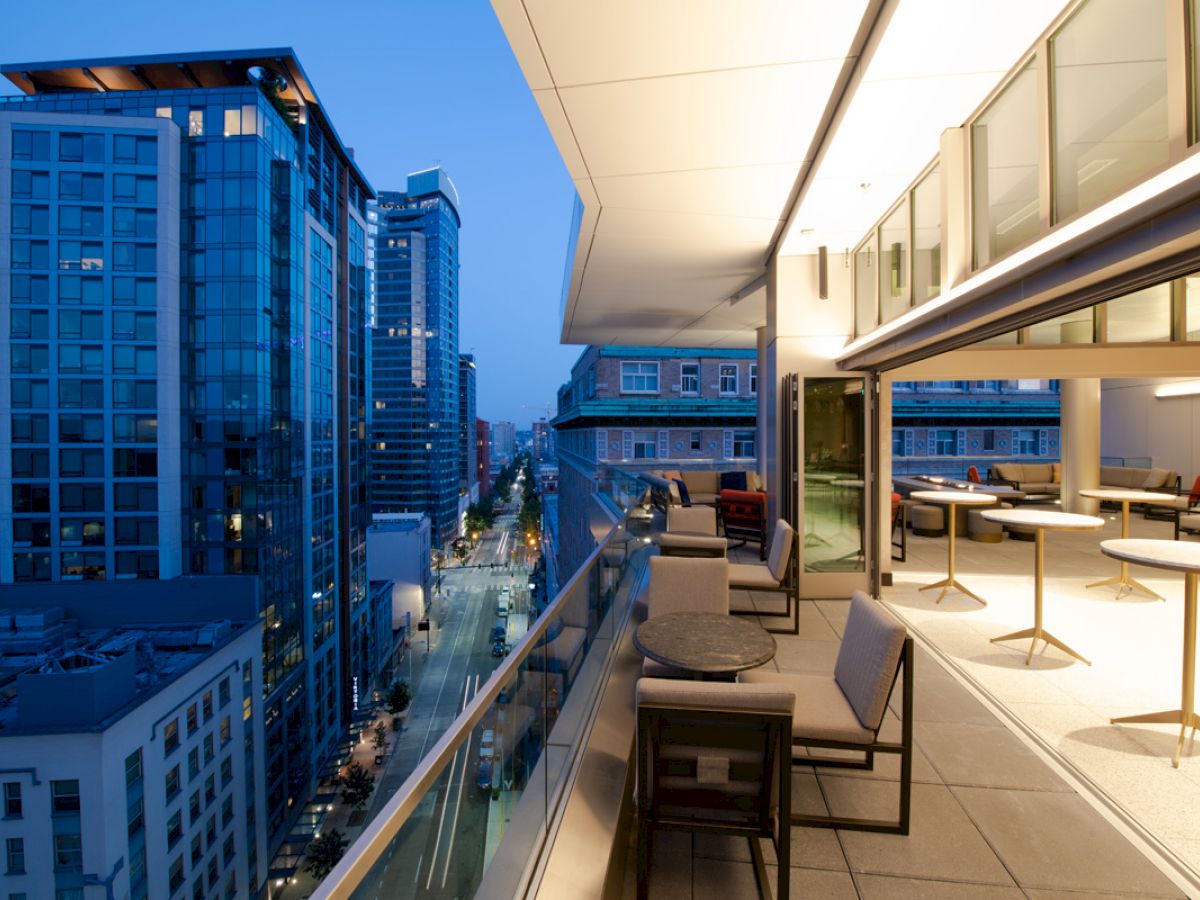 The image shows a modern cityscape at dusk with a balcony view over a busy street, featuring buildings and outdoor seating.